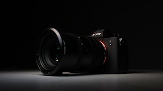 A high-quality camera with a large lens is set against a dark background. The camera is positioned at an angle, with the lens prominently in focus. The overall lighting is low, highlighting the camera's sleek design and details on the lens and body.