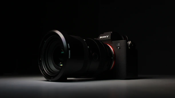 A high-quality camera with a large lens is set against a dark background. The camera is positioned at an angle, with the lens prominently in focus. The overall lighting is low, highlighting the camera's sleek design and details on the lens and body.