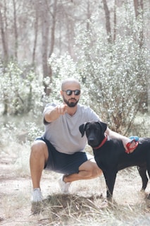 A veteran crouched in a sunlit forest clearing, checking gear while his attentive service dog waits nearby.