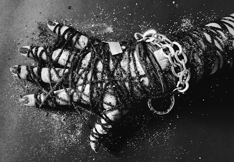 A hand is wrapped in dark cords and metallic chains against a dark background. The fingers and wrist are tightly bound, with glitter sprinkled across the hand and surface. The monochrome color scheme adds depth and contrast to the textures.