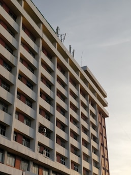 A perspective view of a multi-story building with a series of identical balconies and windows. The structure features a uniform pattern with balconies separated by vertical columns and horizontal beams. Communication antennas are visible on the rooftop under a clear sky.