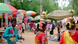 A vibrant outdoor market scene with colorful umbrellas and people milling around. Two women in traditional attire with pots on their heads, decorated with flowers, are in the foreground. A man is playing a drum nearby.