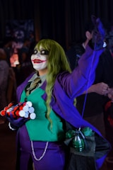 The image features a person dressed in a costume that resembles a comic book villain, with green hair, a purple suit, and green vest. Their face is painted with white makeup, dark eyes, and a large red smile. They hold cylindrical objects resembling explosives and a green grenade is visible attached to their costume.