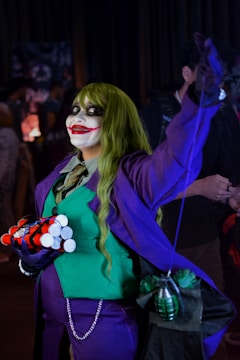 The image features a person dressed in a costume that resembles a comic book villain, with green hair, a purple suit, and green vest. Their face is painted with white makeup, dark eyes, and a large red smile. They hold cylindrical objects resembling explosives and a green grenade is visible attached to their costume.
