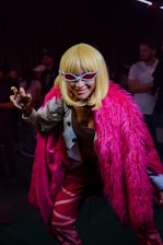 A person is dressed in a striking costume featuring a bright pink faux fur coat and vibrant, patterned pants. They wear a blonde wig and bold, angular sunglasses, striking a playful, claw-like pose. The background is dimly lit with a few people visible, creating a lively, nightclub atmosphere.