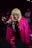 A person is dressed in a striking costume featuring a bright pink faux fur coat and vibrant, patterned pants. They wear a blonde wig and bold, angular sunglasses, striking a playful, claw-like pose. The background is dimly lit with a few people visible, creating a lively, nightclub atmosphere.