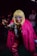 A person is dressed in a striking costume featuring a bright pink faux fur coat and vibrant, patterned pants. They wear a blonde wig and bold, angular sunglasses, striking a playful, claw-like pose. The background is dimly lit with a few people visible, creating a lively, nightclub atmosphere.