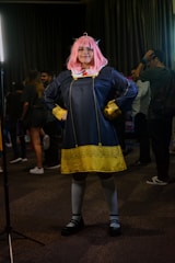 A person is dressed in a cosplay costume, featuring a pink wig and a navy blue dress with yellow details, posing confidently with their hands on their hips. The setting appears to be an indoor event, with other people in casual attire in the background.