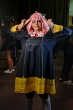 A person in a cosplay costume with pink hair styled with bangs. The costume features a long dark coat with yellow decorative patterns along the hem and cuffs, and a red tie under a white collar. The individual is posing with both hands raised toward their head, fingers forming a gesture.