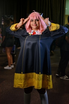 A person in a cosplay costume with pink hair styled with bangs. The costume features a long dark coat with yellow decorative patterns along the hem and cuffs, and a red tie under a white collar. The individual is posing with both hands raised toward their head, fingers forming a gesture.