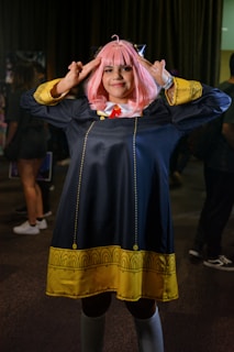 A person in a cosplay costume with pink hair styled with bangs. The costume features a long dark coat with yellow decorative patterns along the hem and cuffs, and a red tie under a white collar. The individual is posing with both hands raised toward their head, fingers forming a gesture.