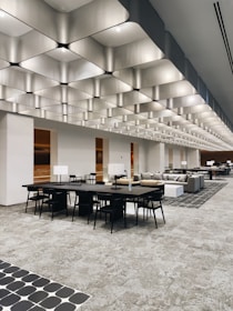 A spacious, modern interior with a striking geometric ceiling design. Multiple black tables and chairs are arranged throughout the open space. Sofas and upholstered seating areas are also present. The lighting is ample, providing a bright ambiance, and there are decorative elements like monochromatic patterns on the floor tiles.