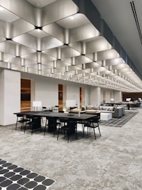 A spacious, modern interior with a striking geometric ceiling design. Multiple black tables and chairs are arranged throughout the open space. Sofas and upholstered seating areas are also present. The lighting is ample, providing a bright ambiance, and there are decorative elements like monochromatic patterns on the floor tiles.