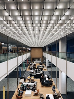 A large, modern interior space with an impressive geometric ceiling of illuminated panels. Below, a spacious open area features several groups of people seated and engaged in conversation. There are tables, chairs, and banners visible in what appears to be a professional or social gathering. The overall layout is symmetrical with a central aisle flanked by elevated walkways.