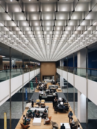 A large, modern interior space with an impressive geometric ceiling of illuminated panels. Below, a spacious open area features several groups of people seated and engaged in conversation. There are tables, chairs, and banners visible in what appears to be a professional or social gathering. The overall layout is symmetrical with a central aisle flanked by elevated walkways.