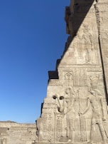 Ancient stone carvings depict figures and hieroglyphics on a wall, set against a clear blue sky. The carvings include detailed representations of human-like figures and intricate symbols.