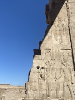 Ancient stone carvings depict figures and hieroglyphics on a wall, set against a clear blue sky. The carvings include detailed representations of human-like figures and intricate symbols.