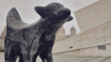 A black sculpture of an abstract animal, covered in white chalk-like drawings, stands prominently in an urban setting. The background features modern architecture with beige and light gray tones and blurred historic buildings in the distance.