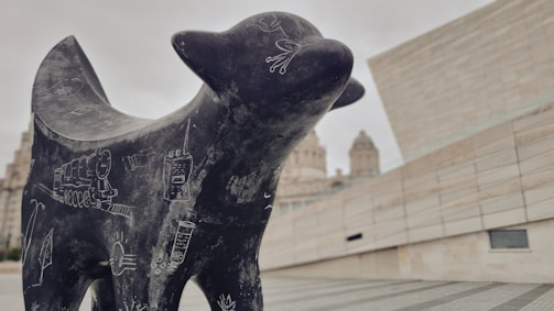 A black sculpture of an abstract animal, covered in white chalk-like drawings, stands prominently in an urban setting. The background features modern architecture with beige and light gray tones and blurred historic buildings in the distance.