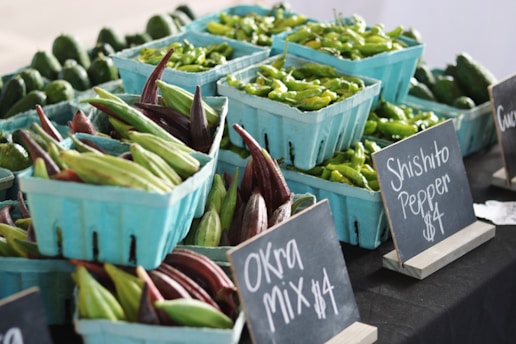 Baskets of fresh green and burgundy okra, along with shishito peppers, are displayed at a market stall. Chalkboard signs in small wooden stands indicate prices for each item. The produce appears fresh and vibrant, arranged neatly in blue containers.
