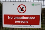 A warning sign is mounted on a wire fence with bold red and white colors, stating 'No unauthorised persons'. There is a prohibition symbol featuring a red circle with a slash over a hand. The logo of the Environment Agency is displayed in the top right corner.