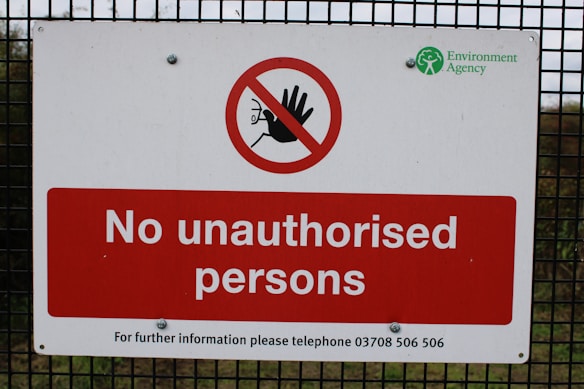 A warning sign is mounted on a wire fence with bold red and white colors, stating 'No unauthorised persons'. There is a prohibition symbol featuring a red circle with a slash over a hand. The logo of the Environment Agency is displayed in the top right corner.