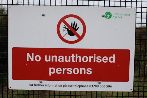 A warning sign is mounted on a wire fence with bold red and white colors, stating 'No unauthorised persons'. There is a prohibition symbol featuring a red circle with a slash over a hand. The logo of the Environment Agency is displayed in the top right corner.