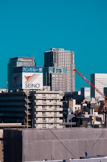 A cityscape with several tall buildings under a bright blue sky. Prominent signage is visible with Japanese characters and a kangaroo logo. A red crane is positioned among the buildings, suggesting ongoing construction. The scene combines both modern and older architectural styles, showcasing residential and commercial structures.
