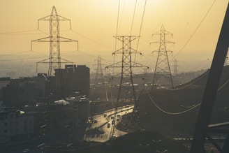 Several high-voltage power lines and transmission towers are seen against a cityscape under a hazy, yellow-tinged sky. Buildings and a winding road with vehicles are visible, providing a sense of urbanization and infrastructure.