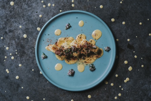 A blue plate with a gourmet dish artfully presented, featuring what appears to be seasoned chicken, creamy sauce, and small garnishes. The dish is styled with additional sauce drizzled around it, and the background is a textured dark surface with random sauce splatters.