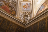 The image displays a richly decorated ceiling corner with intricate artworks and gilded details. There are two small cherub statues facing each other, and multiple fresco paintings, including a central one featuring a cherub playing a violin and others depicting historical or religious scenes. The overall decor includes ornate stucco and gold trim, characteristic of Renaissance or Baroque architecture.