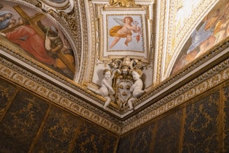 The image displays a richly decorated ceiling corner with intricate artworks and gilded details. There are two small cherub statues facing each other, and multiple fresco paintings, including a central one featuring a cherub playing a violin and others depicting historical or religious scenes. The overall decor includes ornate stucco and gold trim, characteristic of Renaissance or Baroque architecture.