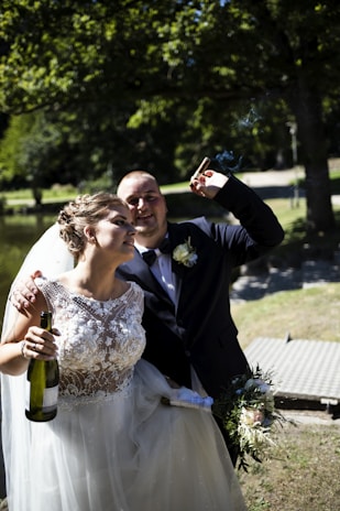 A couple dressed in wedding attire is standing outdoors. The woman is in an elegant, detailed white wedding gown holding a champagne bottle, and the man, wearing a suit with a boutonniere, is smiling and holding a cigar. They are in a park with lush greenery and a pathway in the background.