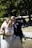 A couple dressed in wedding attire is standing outdoors. The woman is in an elegant, detailed white wedding gown holding a champagne bottle, and the man, wearing a suit with a boutonniere, is smiling and holding a cigar. They are in a park with lush greenery and a pathway in the background.