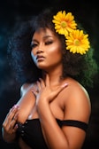 A woman with dark curly hair adorned with two large yellow flowers. She is wearing a black off-shoulder top and has her hands gracefully placed on her chest. Her makeup is elegantly done with highlighted cheekbones and glossy lips. The background is dark, with a soft texture.