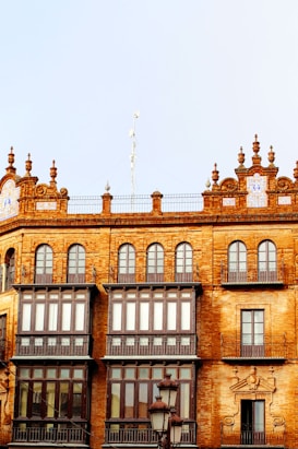 A historic building with intricate architectural details features arched windows and ornate decorations on the rooftop. The structure is made of red-brown bricks, and there are large glass windows with dark wooden frames. Decorative elements include balustrades and artistic carvings. In front of the building, there is a vintage streetlamp.
