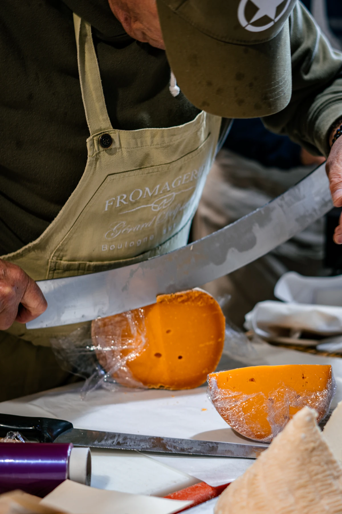 A cheesemonger in a Fromagerie apron slicing aged cheese with a large knife — proper cheese cutting technique