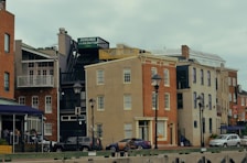 Buildings with a mix of architectural styles line a street, featuring façades in red and beige brick, with various windows and balconies. Two lampposts are visible in the foreground alongside parked cars and a person sitting on a bench. A sign reads 'AVAILABLE' above a staircase on one building.