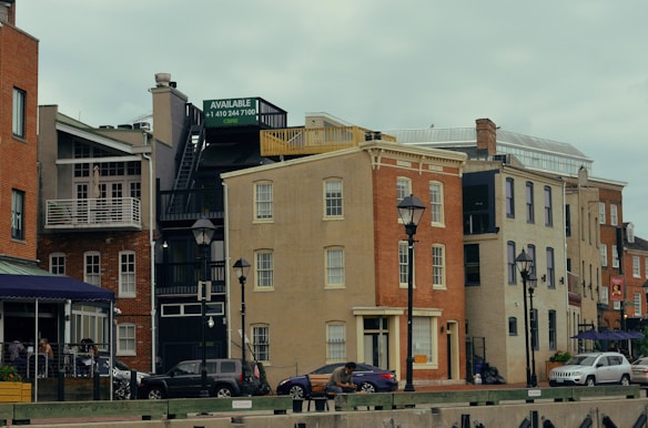 Buildings with a mix of architectural styles line a street, featuring fa&ccedil;ades in red and beige brick, with various windows and balconies. Two lampposts are visible in the foreground alongside parked cars and a person sitting on a bench. A sign reads 'AVAILABLE' above a staircase on one building.