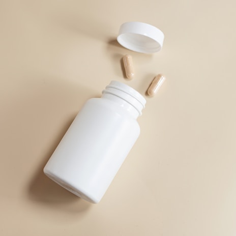 A white plastic pill bottle lies on its side with the cap removed. Two beige capsules are positioned nearby on a pale, neutral-colored surface.