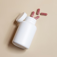 A white plastic bottle lies on its side with four red capsules scattered nearby, all set against a plain beige background. The bottle cap is open, suggesting recent use or preparation.