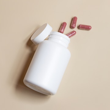 A white plastic bottle lies on its side with four red capsules scattered nearby, all set against a plain beige background. The bottle cap is open, suggesting recent use or preparation.