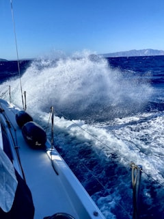 A powerful splash of ocean water is captured as it hits the side of a sailboat, creating a dramatic scene of spray and movement. The deep blue sea extends towards a distant mountainous coastline under a clear blue sky. The boat's deck and safety fenders are visible on the left side.