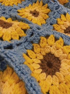 A close-up view of a crochet pattern featuring sunflower designs. The sunflowers consist of yellow petals with dark brown centers, and the background is made up of light blue yarn. The texture of the yarn is prominent, highlighting the intricate weaving.