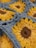 A close-up view of a crochet pattern featuring sunflower designs. The sunflowers consist of yellow petals with dark brown centers, and the background is made up of light blue yarn. The texture of the yarn is prominent, highlighting the intricate weaving.