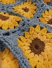 A close-up view of a crochet pattern featuring sunflower designs. The sunflowers consist of yellow petals with dark brown centers, and the background is made up of light blue yarn. The texture of the yarn is prominent, highlighting the intricate weaving.