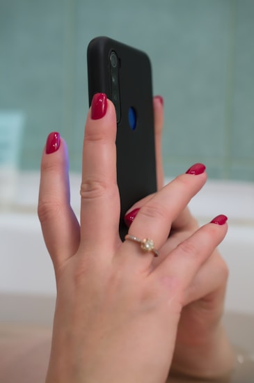 A person holding a smartphone with a black case, displaying well-manicured hands with red nail polish. There is a gold ring with a gemstone on the ring finger. The background appears to be a blurred interior setting with a greenish-blue hue.