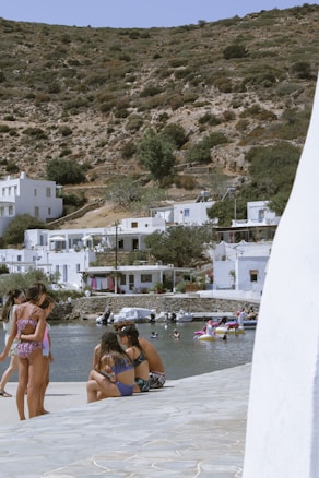 A coastal scene featuring a group of people in swimsuits relaxing and enjoying themselves near the water. The background includes white buildings nestled along the rocky, shrub-covered hillside. Several small boats float in the water, and others are seen swimming or using inflatable rings.