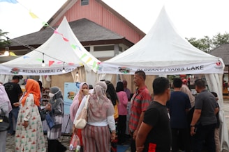 A bustling outdoor market scene features multiple people gathered around white tents with banners advertising food products. Attendees are dressed in a variety of colorful outfits, some wearing headscarves, as they converse and browse the stalls. Decorative streamers are strung between tents, fostering a festive atmosphere.