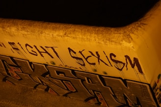 The image depicts graffiti art on a concrete wall. The words 'FIGHT SEXISM' are spray-painted in bold, dark letters, while below them there is another section of graffiti with abstract shapes and letters. The wall and the surrounding area appear weathered and the lighting gives a yellowish hue to the scene.